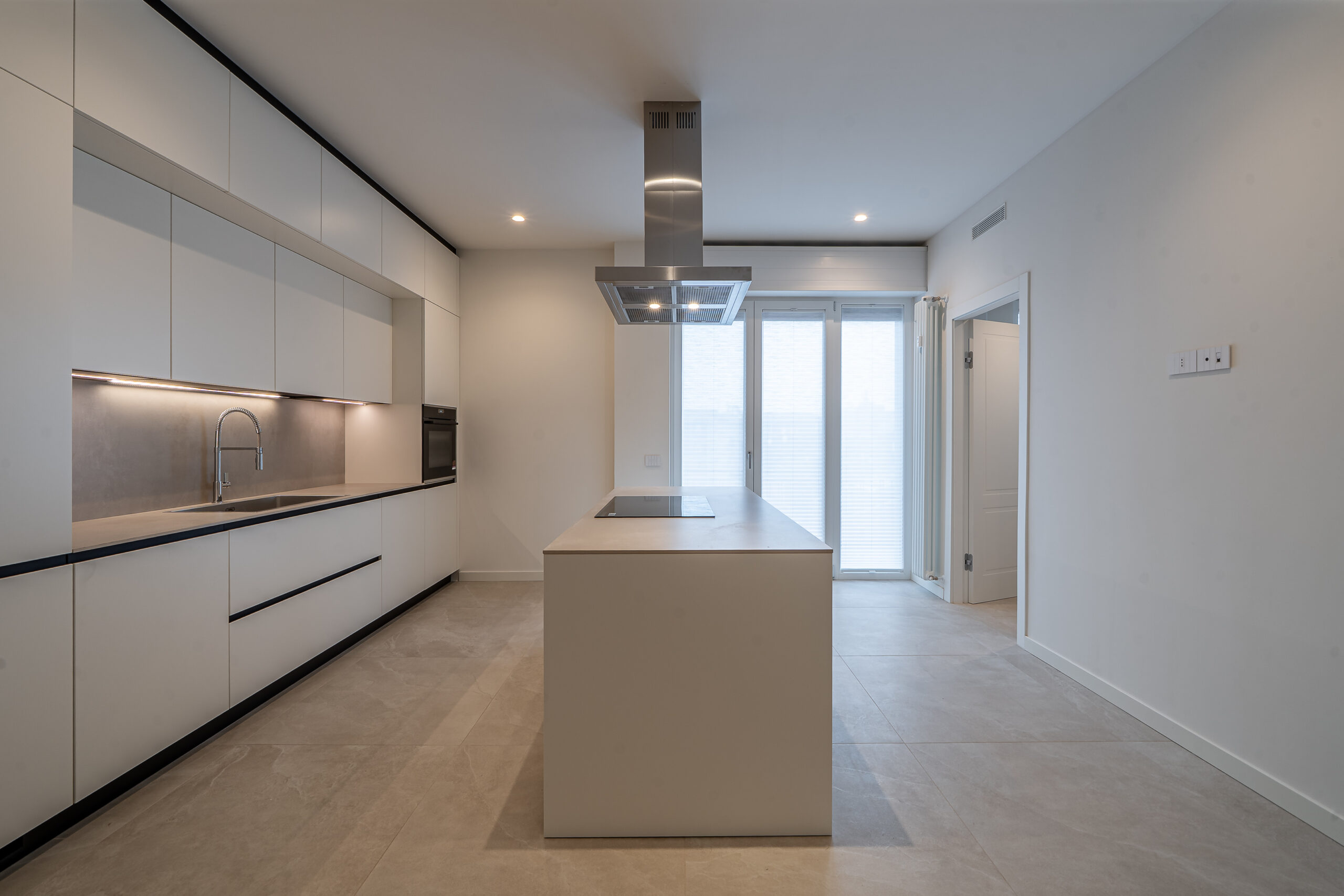 Cucina contemporanea con mobili bianchi, profili neri, isola centrale con piano a induzione e cappa sospesa.